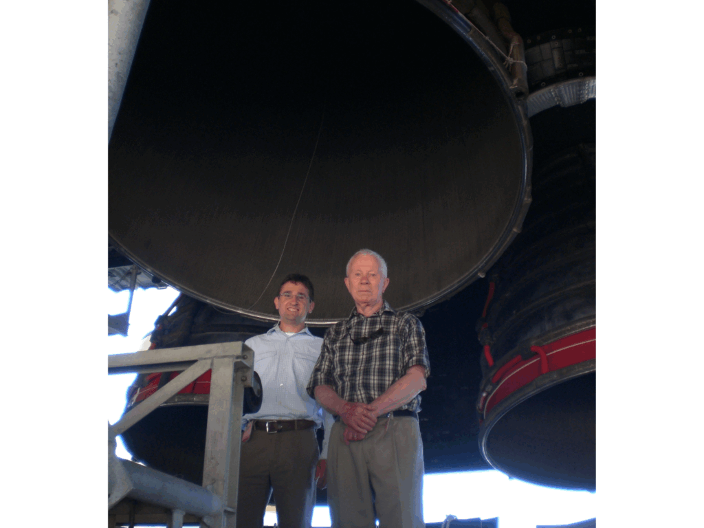 Charlie Danaher In May 2025 Cold Facts: 'A Salute To An Old Friend, Glen Mcintosh' 2 Two men stand beneath the massive nozzles of a rocket engine at nasa's cape canaveral. The man on the left wears a light-colored button-down shirt and smiles at the camera, while the man on the right, older and in a plaid shirt, stands with his hands folded. The towering engine structure above them dominates the background, emphasizing the scale of the aerospace setting.