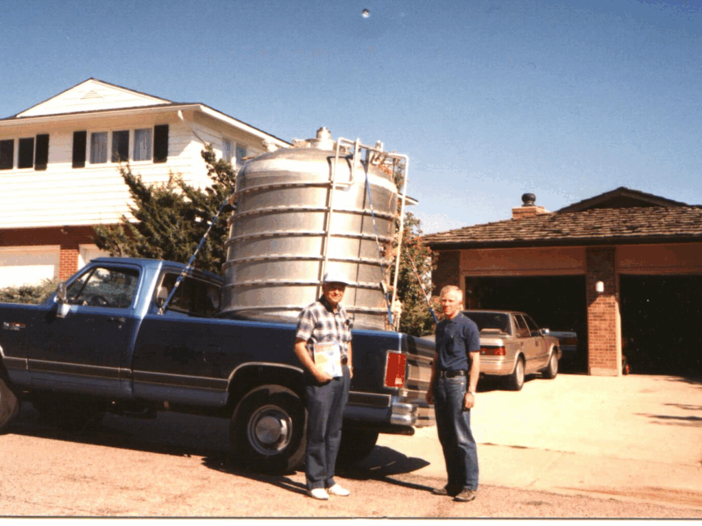 Charlie Danaher In May 2025 Cold Facts: 'A Salute To An Old Friend, Glen Mcintosh' 1 Two men stand beside a dark blue pickup truck parked in front of a suburban house. The truck bed holds a large, cylindrical 750-gallon helium dewar. The man on the left wears a plaid shirt and cap, while the man on the right wears a navy blue shirt and jeans. The scene appears to be from around 1990, with a clear sky overhead and a car parked in a neighboring driveway.